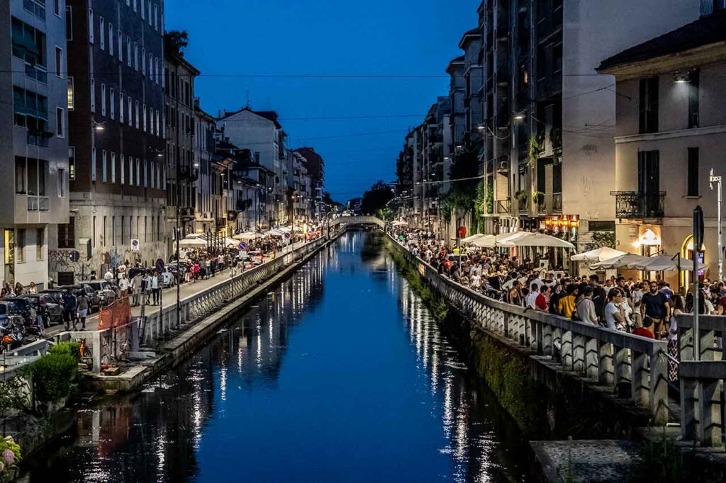 Tętniące życiem nocne życie nad kanałem Naviglio w Mediolanie. Obraz przedstawia tętniącą życiem scenę z oświetlonymi barami, restauracjami i klubami wzdłuż kanału. Ludzie gromadzą się, ciesząc się żywą atmosferą, nawiązując kontakty towarzyskie i tworząc niezapomniane chwile na tle uroczego kanału i jego tętniącego życiem nocnego życia.