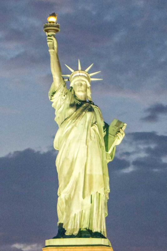 Statua Wolności w Nowym Jorku, oświetlona nocą, widziana z bliskiej perspektywy z pokładu statku.