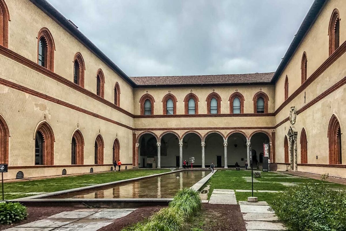 Wewnętrzny dziedziniec Castello Sforzesco w letnie popołudnie, z beżowymi ścianami, arkadami, małym basenem, zieloną trawą i kolorowymi kwiatami pod błękitnym niebem.