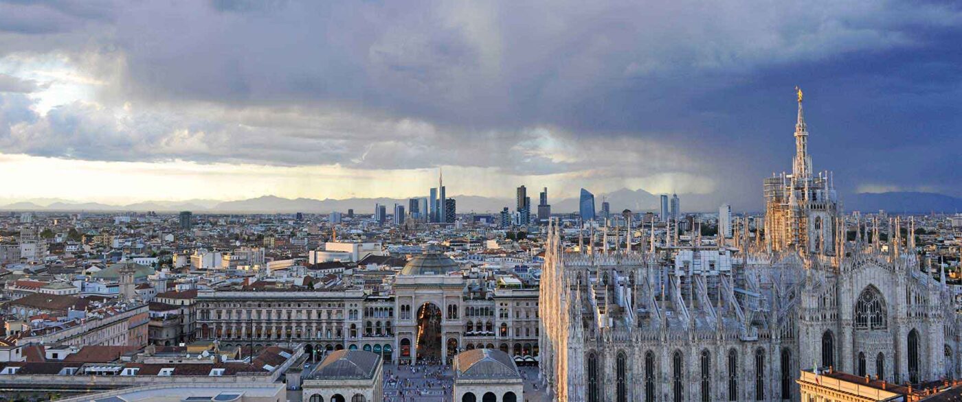 Widok na panoramę Mediolanu o zachodzie słońca, z charakterystycznymi wieżowcami i zabytkowymi budynkami w ciepłym świetle.