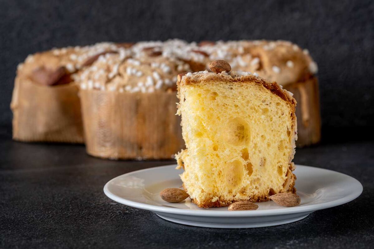 Wielkanoc w Mediolanie. Kawałek colomby pasquale na białym talerzu, w tle całe włoskie ciasto wielkanocne z migdałami i cukrem